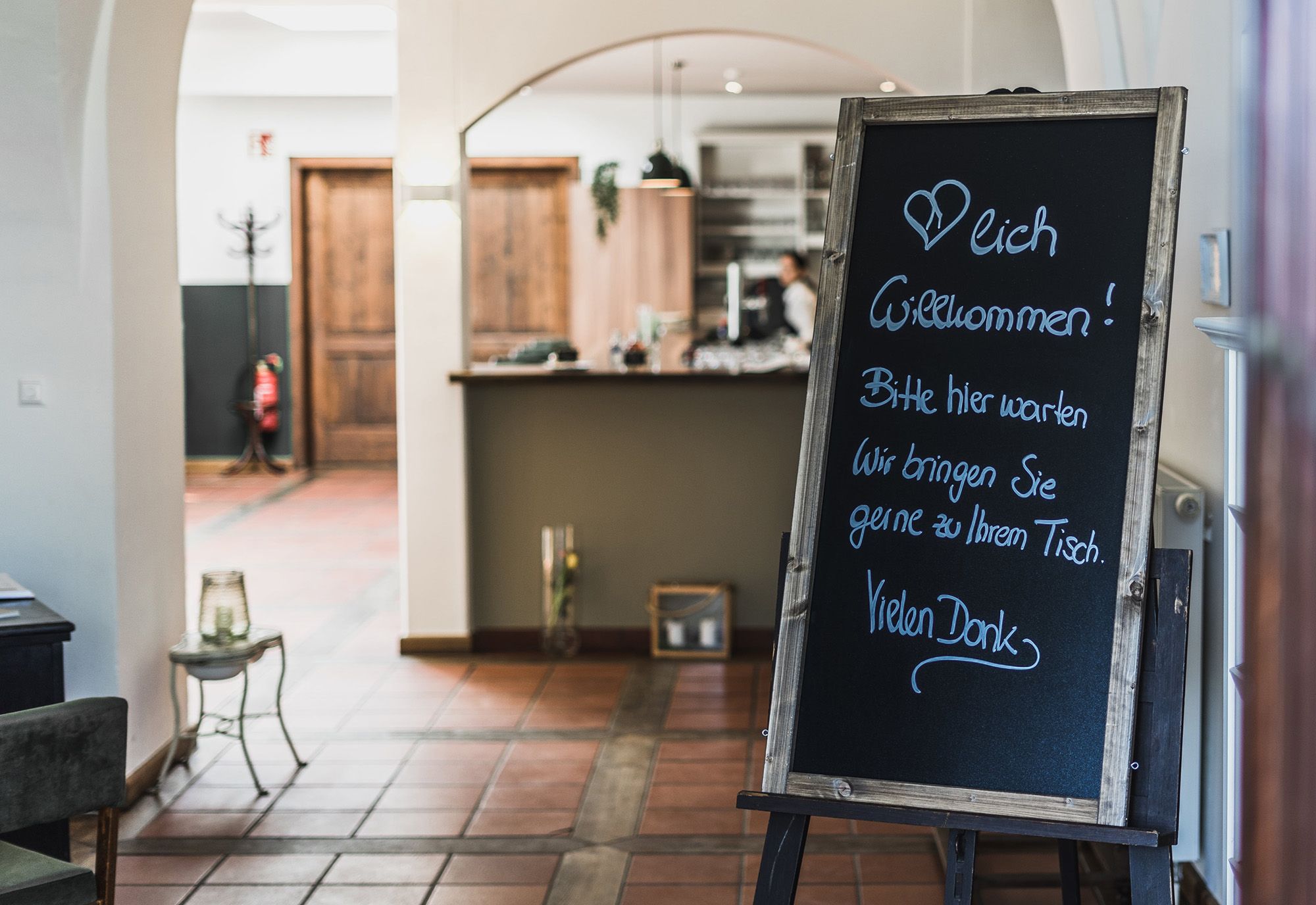 Eingangsbereich eines Restaurants mit einer Tafel, die Gäste willkommen heißt und sie bittet, zu warten, bis sie zu ihrem Tisch gebracht werden.