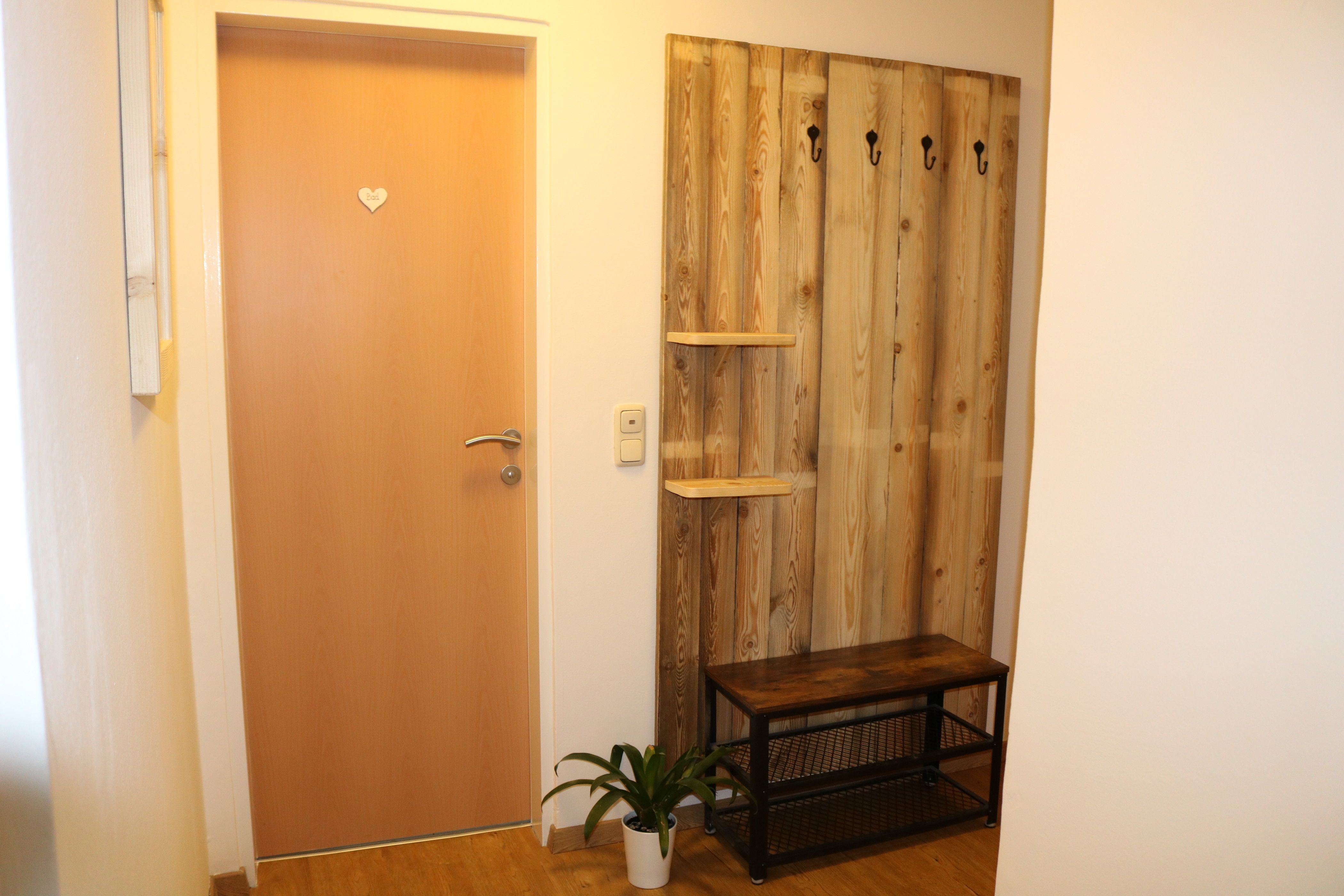Eingangsbereich mit Holztür, Garderobe aus Holz mit Haken, Bank und Pflanze.