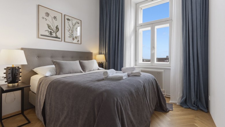 Ein elegantes Schlafzimmer mit einem gro&szlig;en Bett, grauer Bettw&auml;sche, zwei Nachttischen mit Lampen und einem Fenster mit blauem Vorhang.