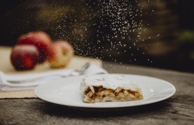 Apfelstrudel mit Puderzucker auf Teller, Äpfel im Hintergrund.