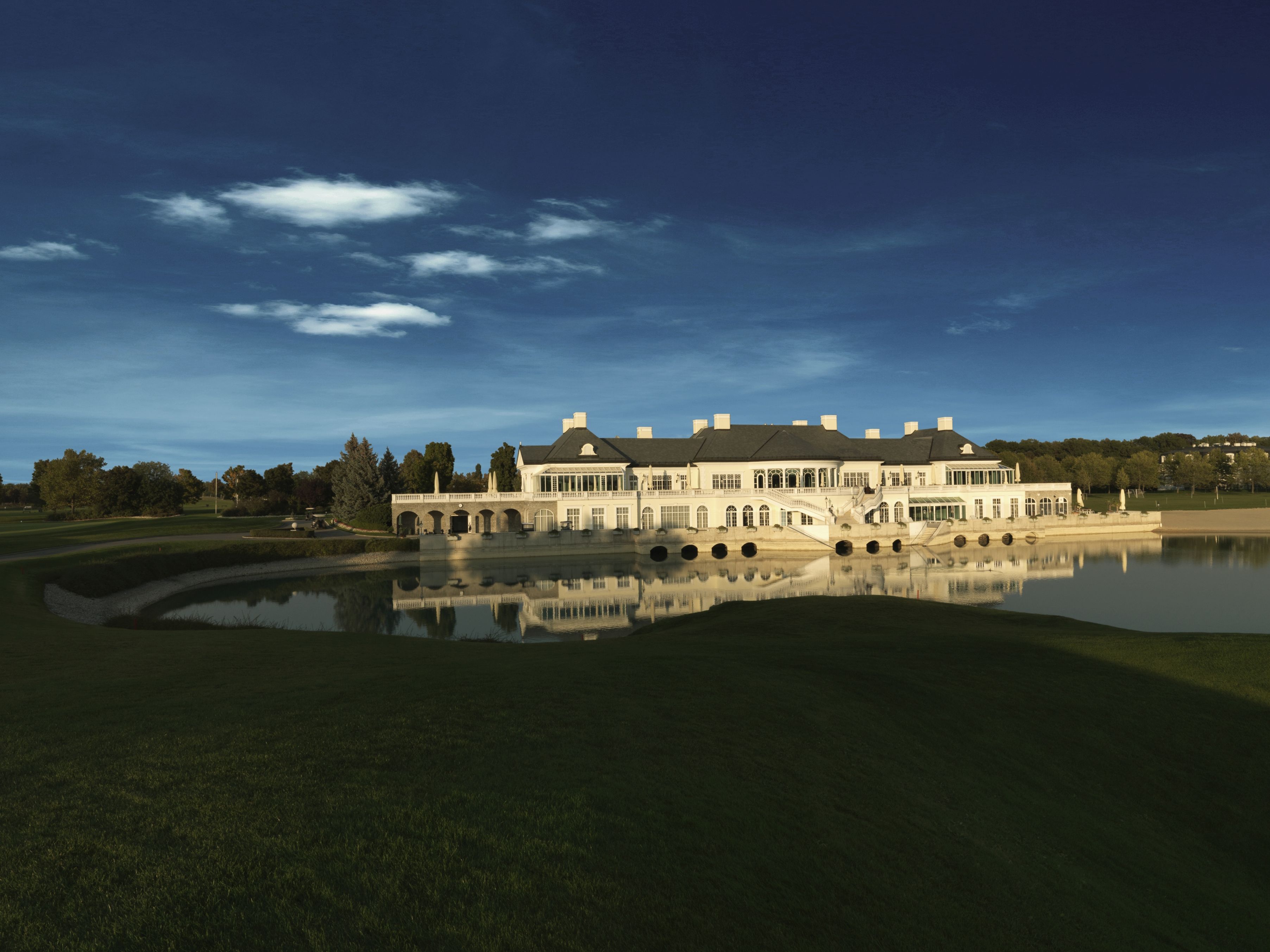 Ein elegantes Clubhaus spiegelt sich in einem ruhigen See, umgeben von grünen Rasenflächen und Bäumen unter einem klaren blauen Himmel.