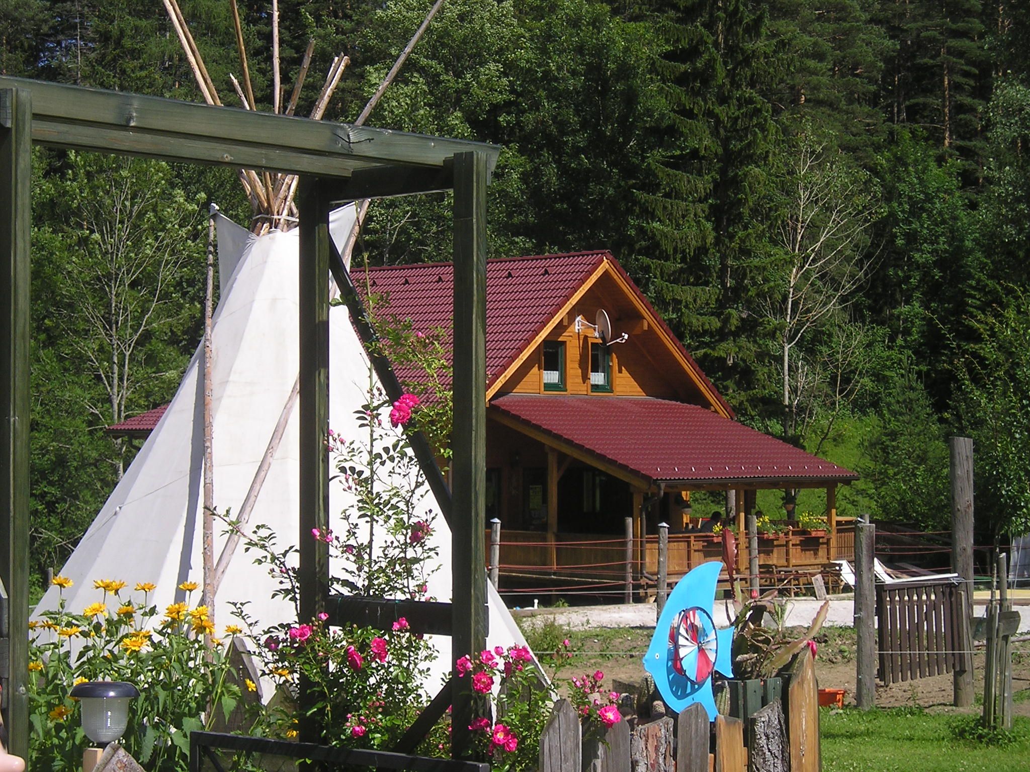 Ein Holzhaus mit rotem Dach, ein Tipi und bunte Blumen im Vordergrund, umgeben von Bäumen.