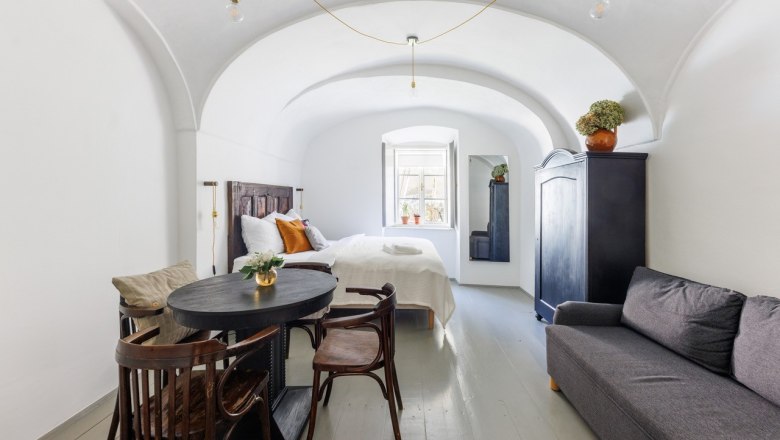 Ein stilvoll eingerichtetes G&auml;stezimmer mit gew&ouml;lbter Decke, Bett, Sofa, Tisch und St&uuml;hlen. Ein Fenster l&auml;sst Tageslicht herein.