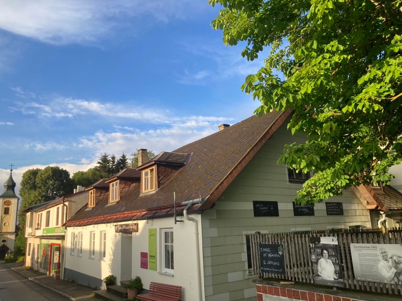 Außenansicht eines traditionellen Wirtshauses mit Schindeldach und grüner Fassade, umgeben von Bäumen und blauem Himmel.