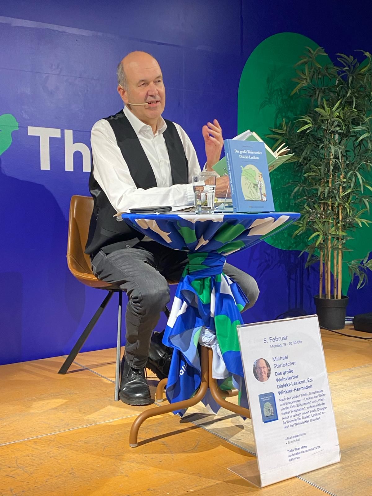 Ein Mann sitzt an einem Tisch und hält eine Lesung. Vor ihm steht ein Buch mit dem Titel 'Das große Wienerler Dialekt-Lexikon'.