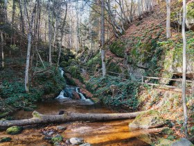 In der herbstlichen Idylle flie&szlig;t ein klarer Bach sanft durch die bewaldete Landschaft, umgeben von bunten Bl&auml;ttern, die in warmen T&ouml;nen leuchten. Die ruhige Atmosph&auml;re l&auml;dt dazu ein, die frische Luft zu genie&szlig;en und die Sch&ouml;nheit der Natur zu erkunden.