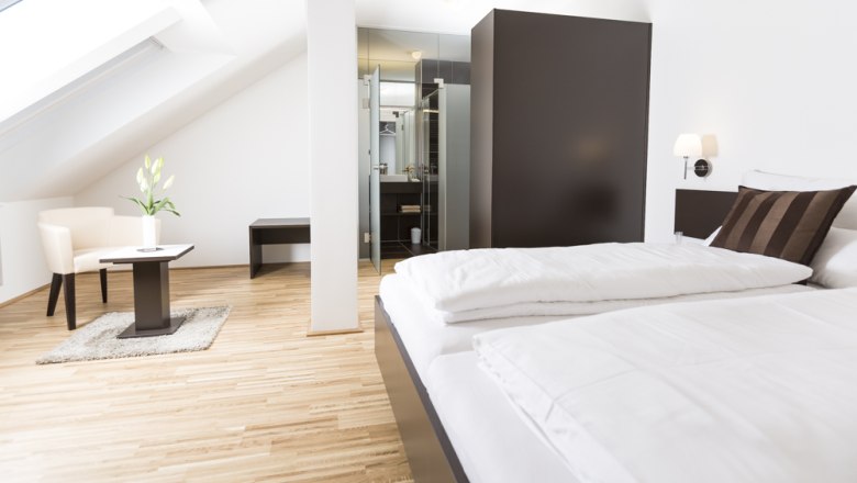 Gästezimmer, © Ernst Jöchlinger Modernes Gästezimmer mit Bett, Sessel und Tisch unter Dachschräge.