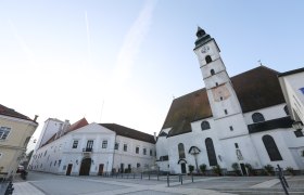 Pfarrkirche mit Uhrturm und angrenzenden Geb&auml;uden auf einem leeren Platz.