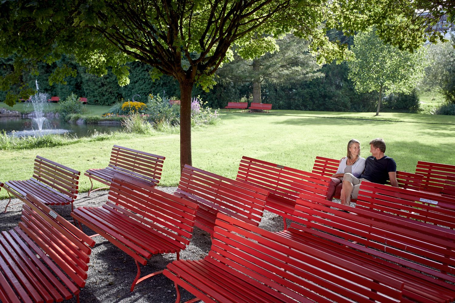 Ein Paar sitzt auf roten Bänken im Kurpark, umgeben von Bäumen und einem Teich mit Springbrunnen.