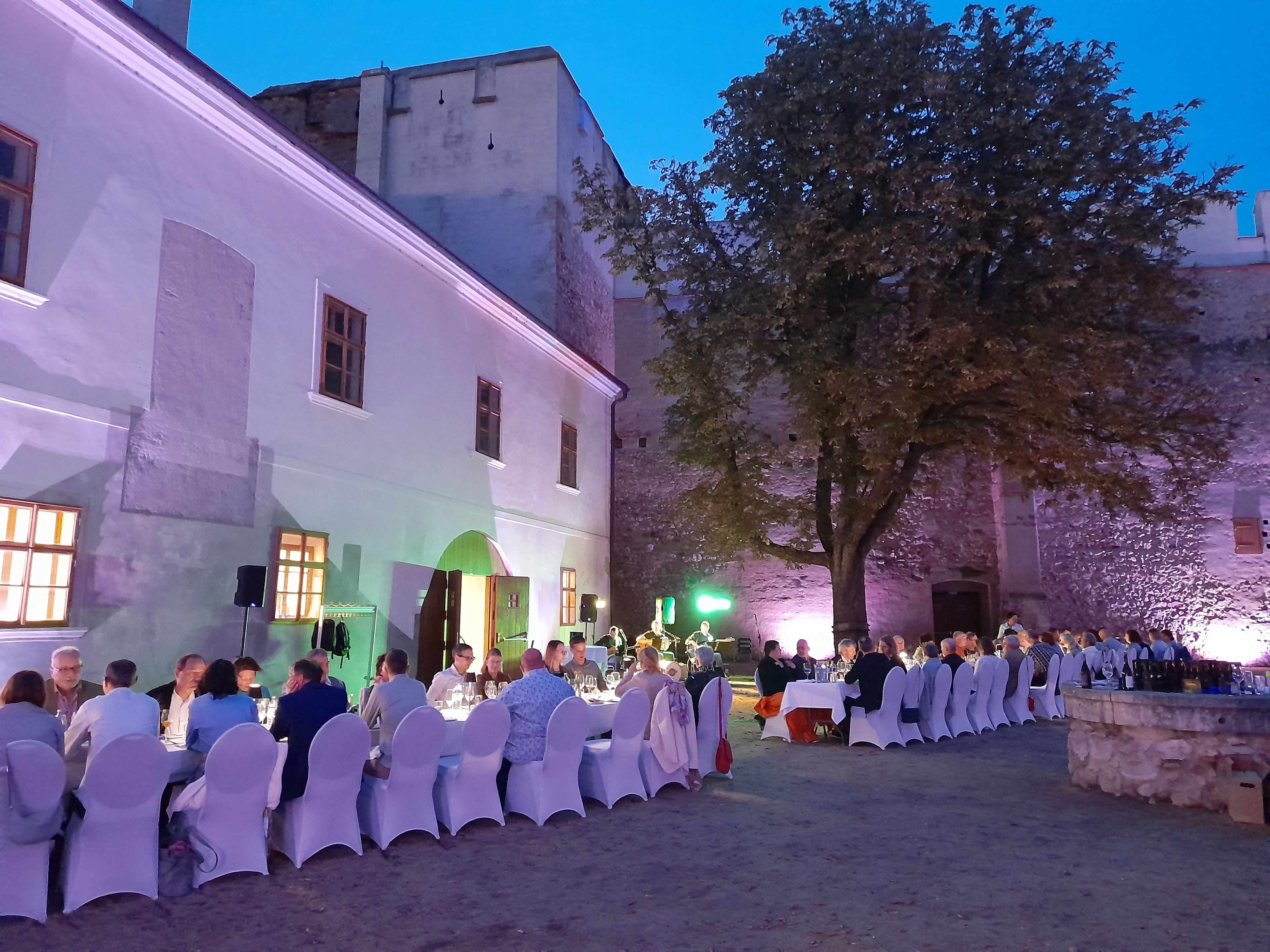 Abendveranstaltung im Freien mit gedeckten Tischen und Gästen vor einer beleuchteten historischen Mauer.