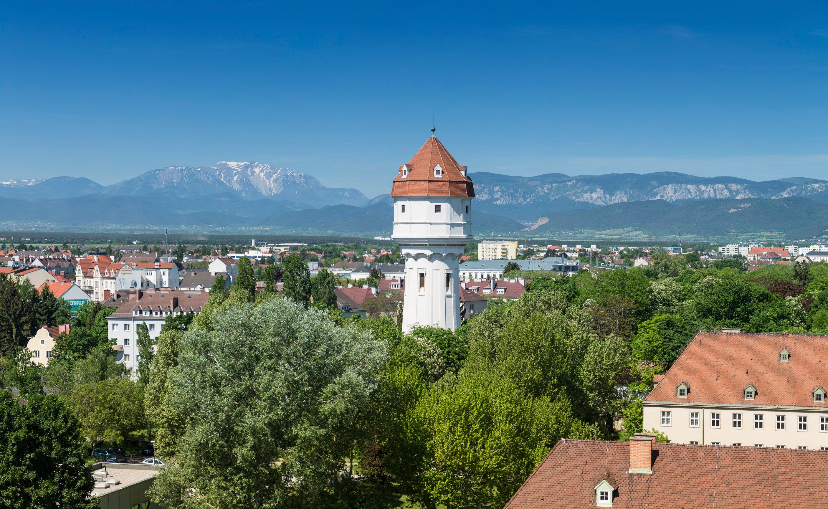 Ein weißer Wasserturm mit rotem Dach in einer grünen Stadtlandschaft vor einem Bergpanorama.