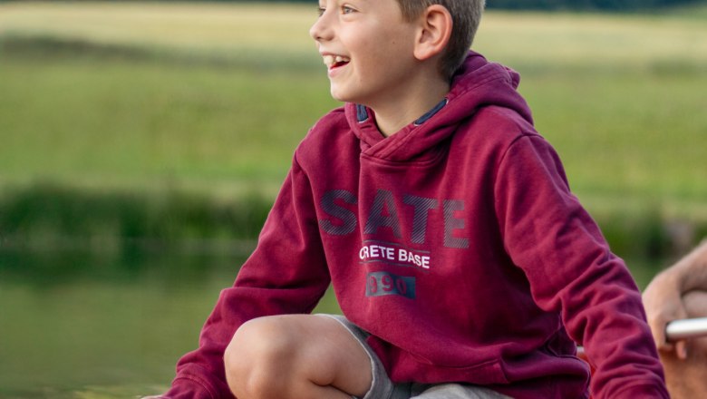 Ein Junge in einem roten Hoodie sitzt lächelnd auf einem Boot auf einem Teich..