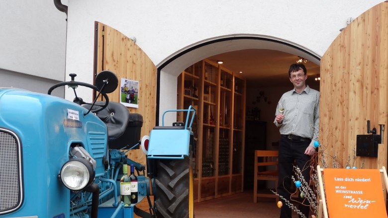Ein Mann steht mit einem Weinglas in der Hand vor einem Geb&auml;ude mit offenen Holzt&uuml;ren. Ein blauer Traktor steht daneben.