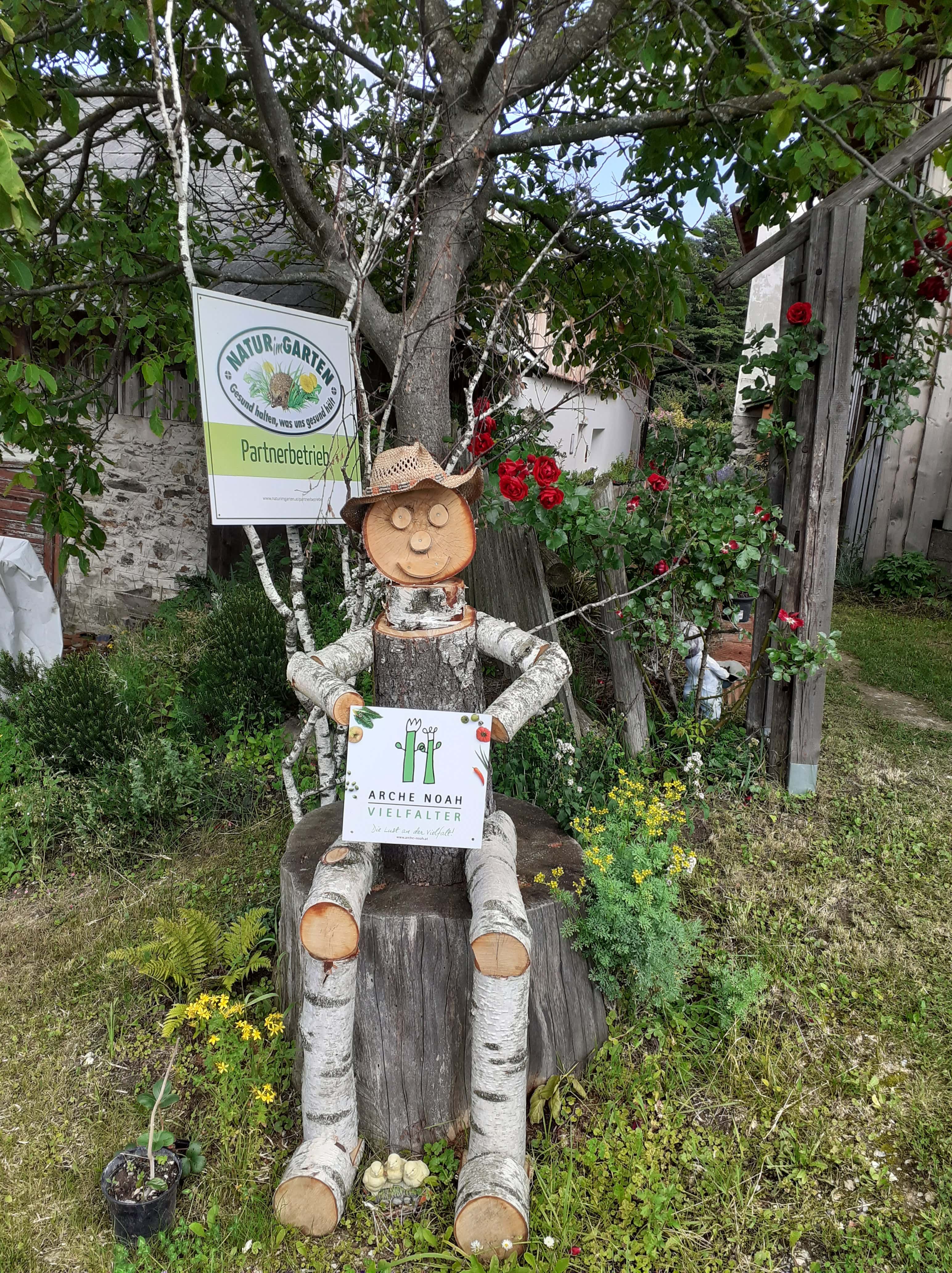 Holzfigur mit Strohhut sitzt auf Baumstumpf, hält Schild mit Aufschrift 'Arche Noah Vielfalter'. Hintergrund: Natur im Garten Schild
