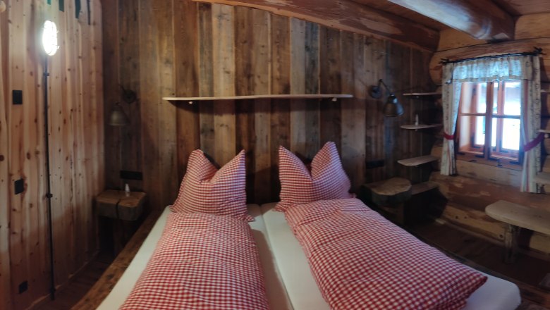 Rustikales Zimmer mit Holzwänden, Bett mit rot-weiß karierten Kissen, Fenster mit Vorhängen, Holztür und Regalen.