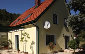 Gelbes Haus mit rotem Dach und Satellitensch&uuml;ssel, umgeben von B&auml;umen.