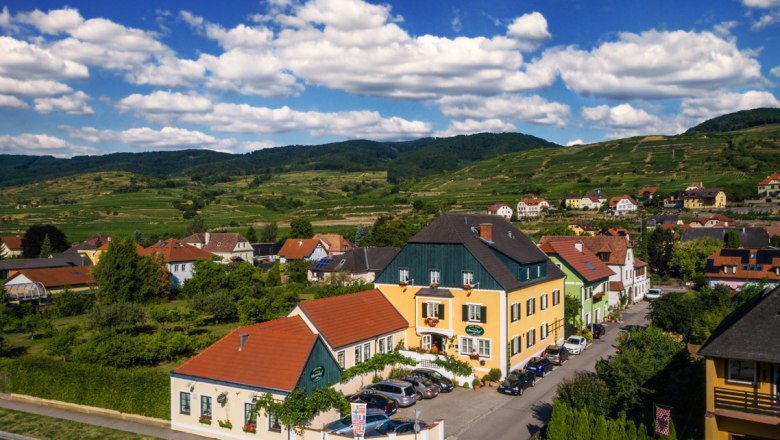 L&auml;ndliche H&auml;user in einer gr&uuml;nen H&uuml;gellandschaft unter blauem Himmel mit Wolken.