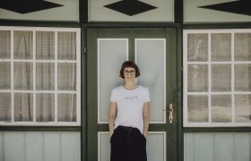 Eine Frau steht vor einer gr&uuml;nen T&uuml;r mit Fenstern und Spitzenvorh&auml;ngen.