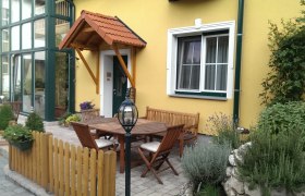 Ein gem&uuml;tlicher Innenhof mit Holzm&ouml;beln, Pflanzen und gelber Hausfassade.