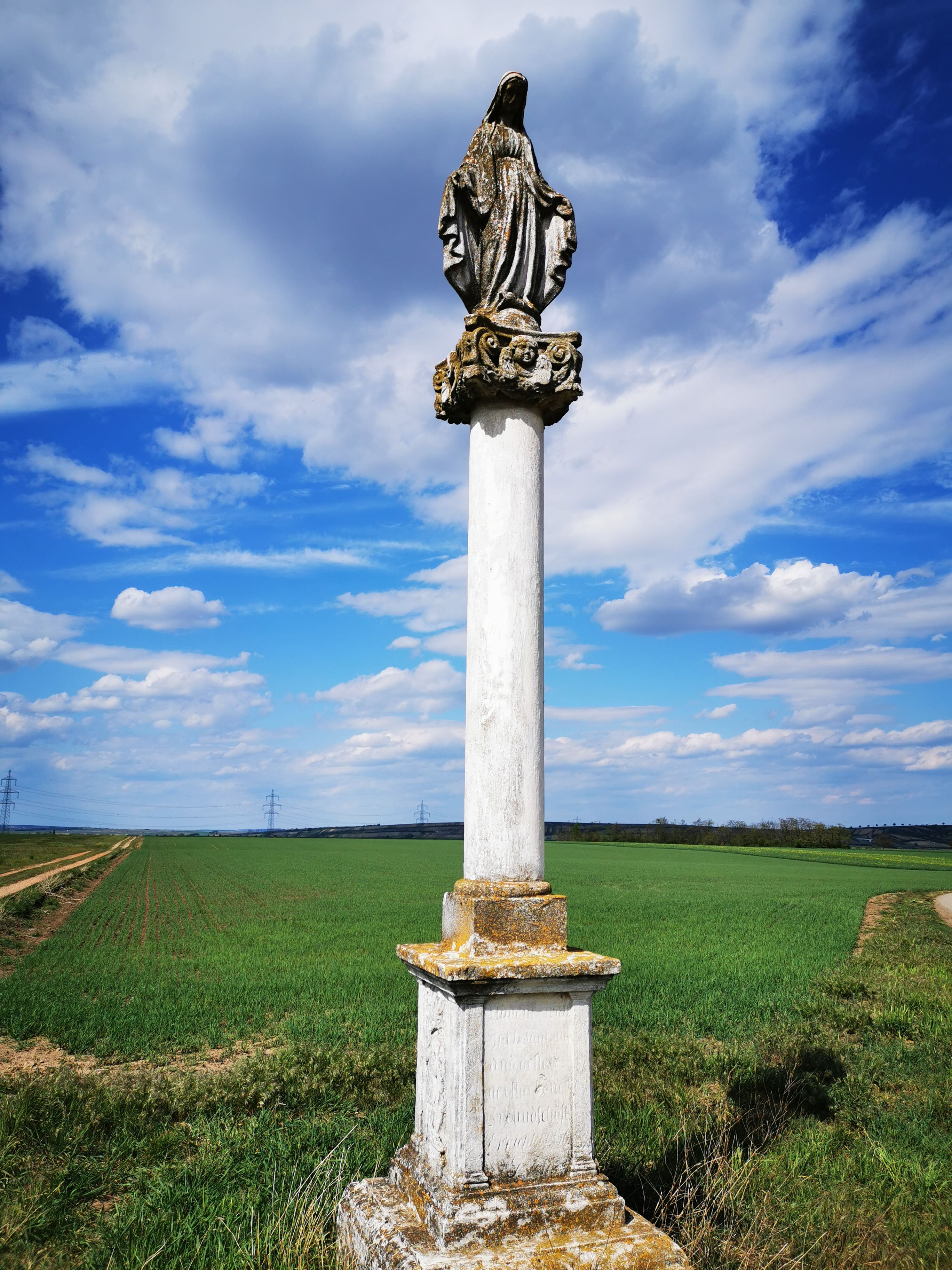 Eine Statue auf einer Säule in einer ländlichen Landschaft unter blauem Himmel mit Wolken.