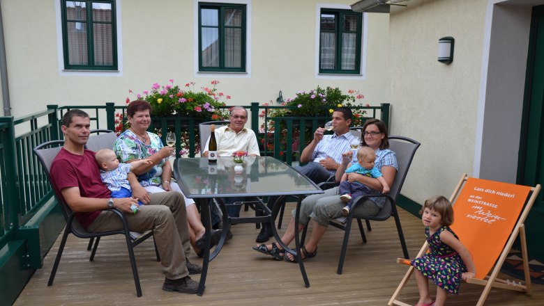 Familie Krottendorfer, © Familie Krottendorfer Eine Familie sitzt auf einer Terrasse an einem Tisch mit Getränken. Im Hintergrund sind Blumen und ein Gebäude zu sehen.