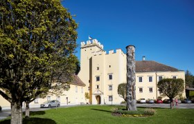 Schloss Gro&szlig;-Siegharts mit gelbem Geb&auml;ude und Turm, umgeben von B&auml;umen und einer Wiese mit Skulptur.