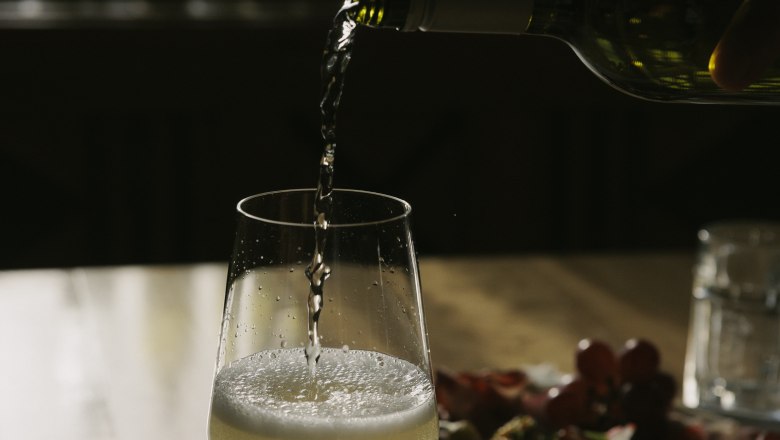 Ein Glas Wei&szlig;wein wird auf einem Tisch eingeschenkt, im Hintergrund ein Teller mit Snacks.
