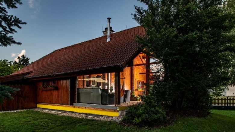 Au&szlig;enansicht eines Holzhauses mit gro&szlig;en Fenstern und Garten, umgeben von B&auml;umen.