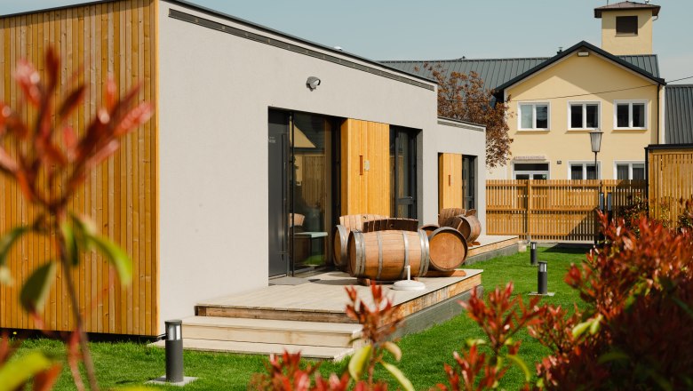 Ipsmiller Appartements, © Michael Reidinger Moderne Appartements mit Holzverkleidung und Weinfässern auf der Terrasse, umgeben von grünem Rasen und einem Zaun.