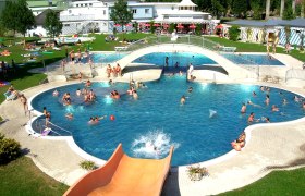 Freibad mit Pool, Brücke und Rutsche, viele Menschen beim Schwimmen und Sonnenbaden.