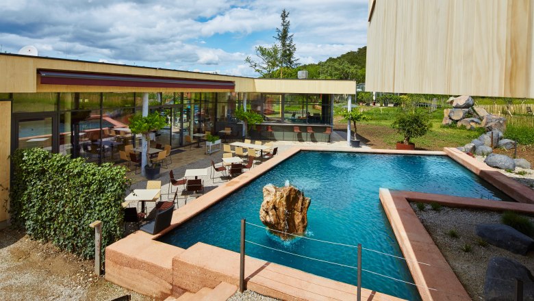 Moderne Terrasse mit Tischen und St&uuml;hlen neben einem rechteckigen Teich mit Felsen in der Mitte.