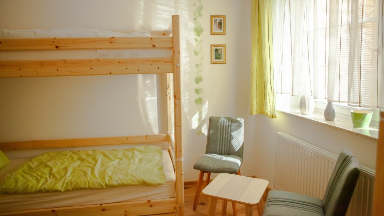 Ein helles Zimmer mit einem Stockbett, zwei St&uuml;hlen, einem kleinen Tisch und einem Fenster mit Vorh&auml;ngen.