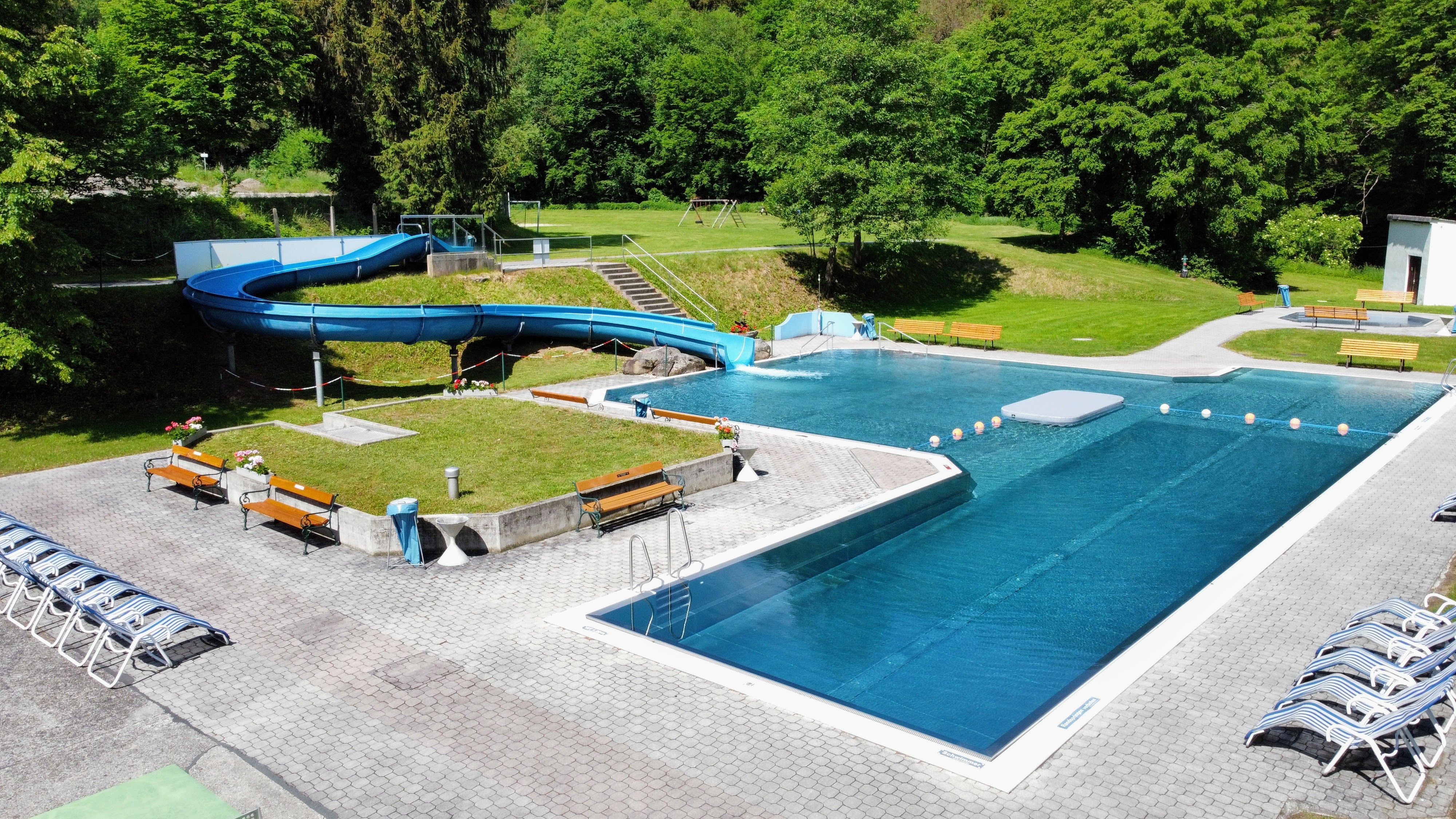 Ein Freibad mit Rutsche und Liegestühlen, umgeben von Bäumen.
