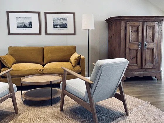 Gemütliches Wohnzimmer mit gelbem Sofa, zwei grauen Sesseln, rundem Holztisch und rustikalem Holzschrank.