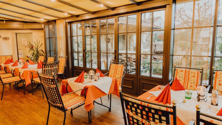 Ein gem&uuml;tlicher Wintergarten mit gedeckten Tischen und St&uuml;hlen, umgeben von gro&szlig;en Fenstern mit Blick nach drau&szlig;en.