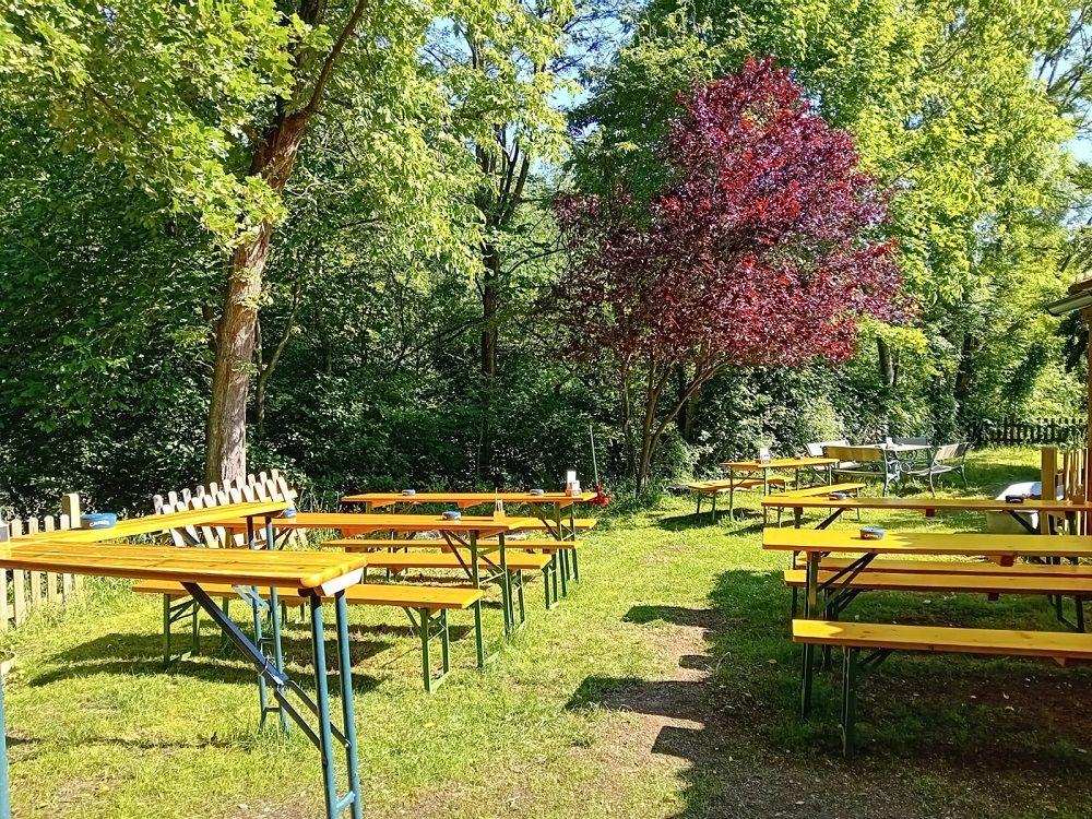 Ein Biergarten mit leeren Holzbänken und Tischen unter Bäumen.