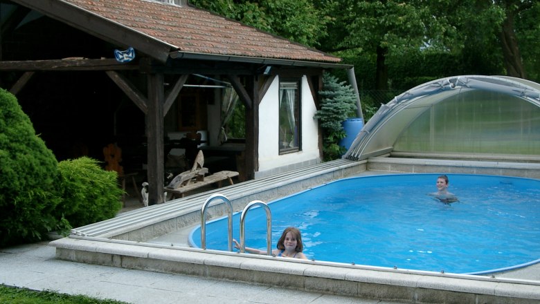 Zwei Kinder schwimmen in einem kleinen, &uuml;berdachten Pool neben einem Holzpavillon.