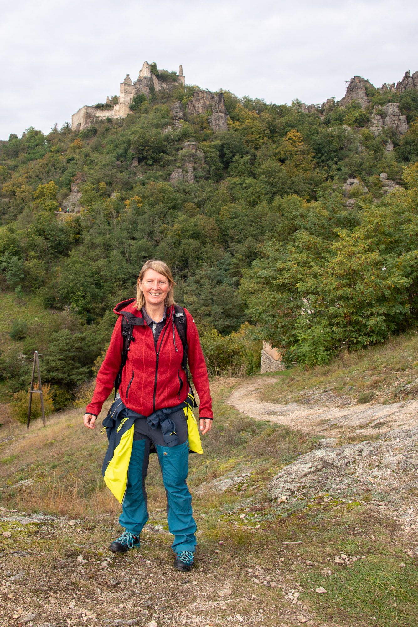 Reiseleiterin Christine Emberger steht lächelnd auf einem Wanderweg mit Blick auf die Ruine Dürnstein im Hintergrund.