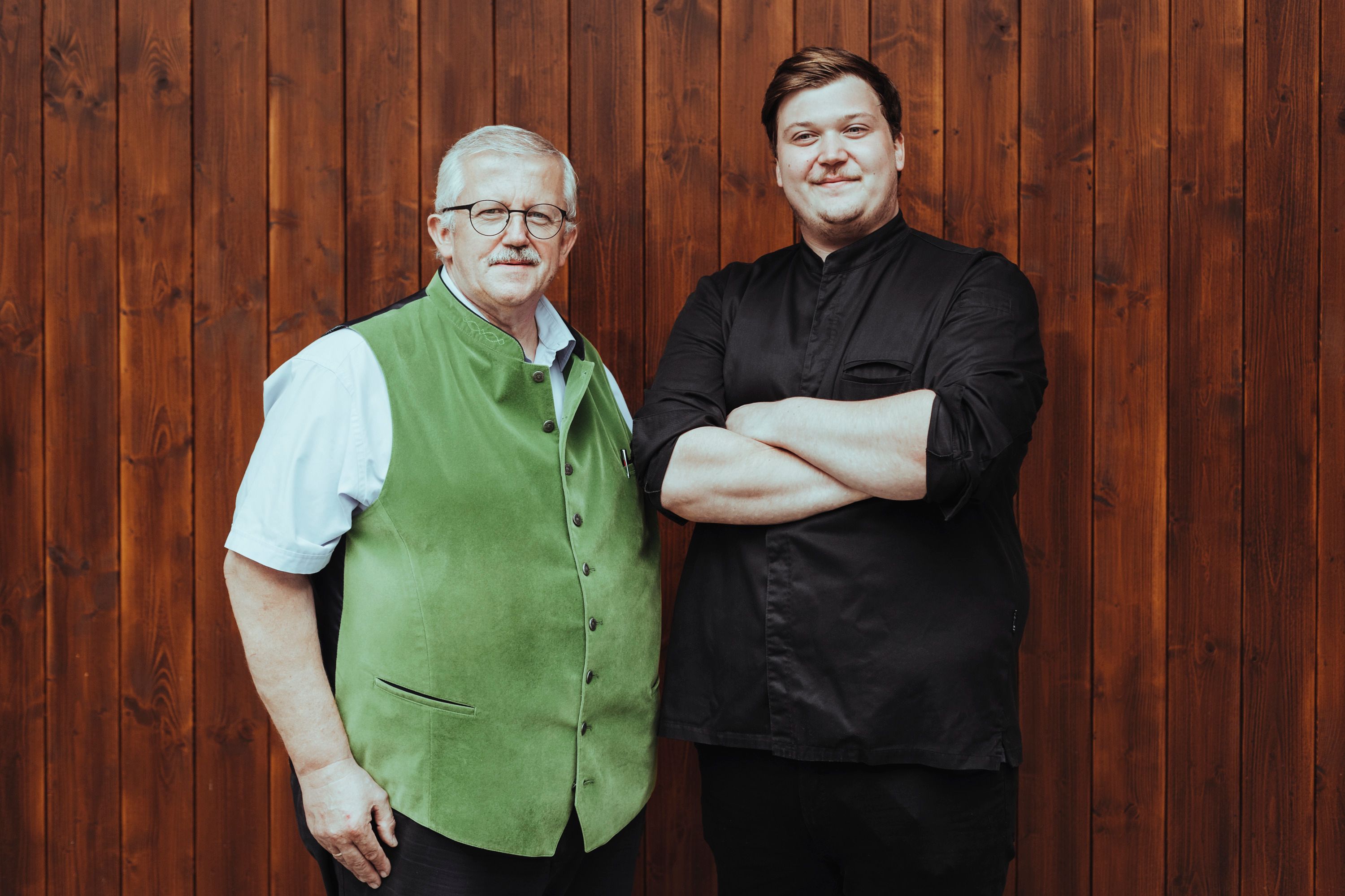 Zwei Männer stehen vor einer Holzwand, einer trägt eine grüne Weste, der andere ein schwarzes Hemd.