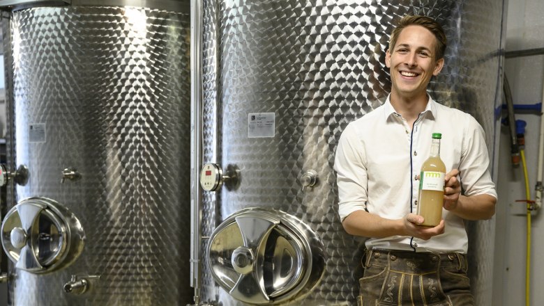 Mann in traditioneller Kleidung h&auml;lt eine Flasche vor gro&szlig;en Metalltanks.