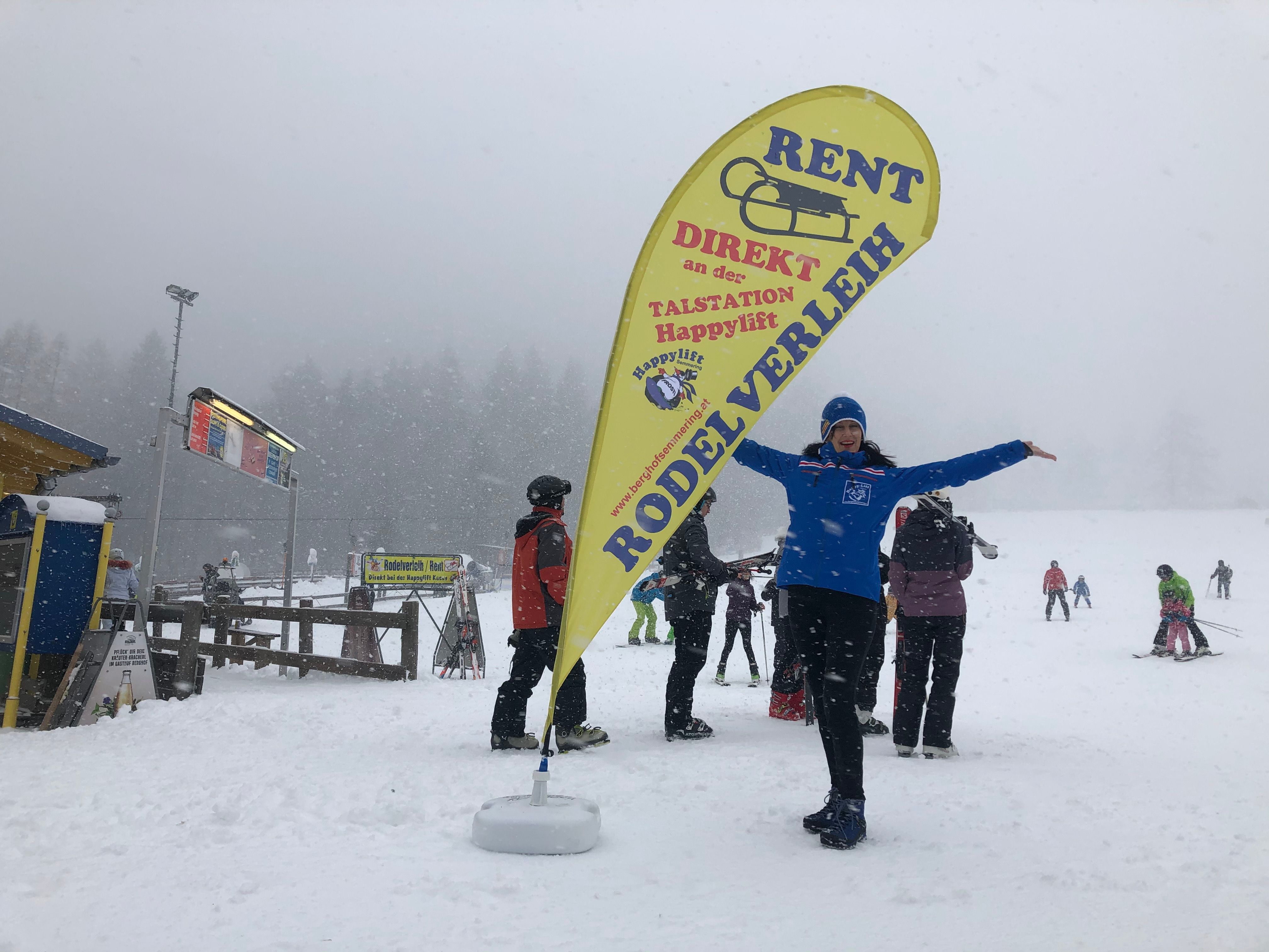 Person im Schnee neben einem gelben Beachflag mit der Aufschrift 'Rodelverleih'.