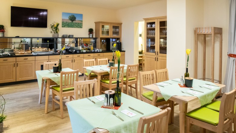 Ein gem&uuml;tlicher Fr&uuml;hst&uuml;cksraum mit Holzm&ouml;beln, gedeckten Tischen und einem Buffetbereich. An den W&auml;nden h&auml;ngen Bilder und Pflanzen dekorieren den Raum.