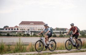 Zwei Radfahrer genie&szlig;en die frische Luft und die malerische Landschaft entlang eines ruhigen Kanals. Die sanften H&uuml;gel im Hintergrund und die &uuml;ppige Vegetation schaffen eine einladende Atmosph&auml;re f&uuml;r Outdoor-Enthusiasten. Hier wird der Sommer in seiner sch&ouml;nsten Form erlebbar.