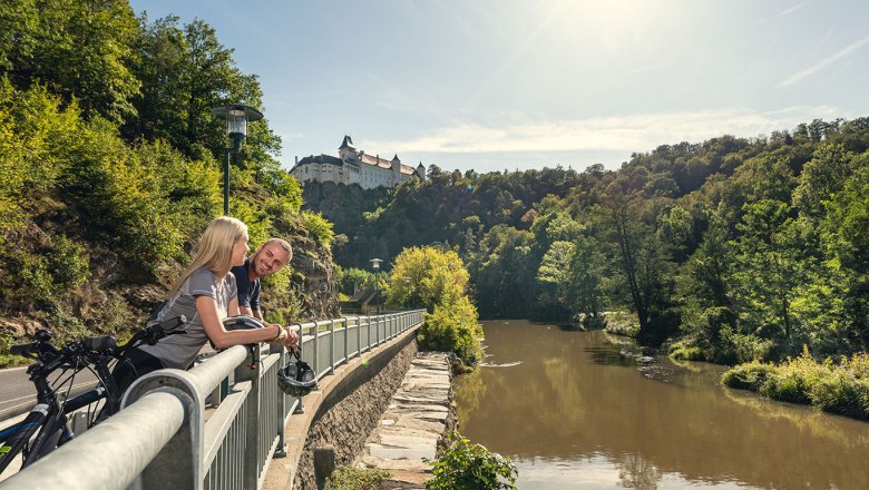 Zwei Personen lehnen an einem Gel&auml;nder mit Blick auf einen Fluss und eine Burg im Hintergrund.
