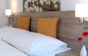 Doppelbett mit Holzr&uuml;ckwand, gelben Kissen und Wandlampen.