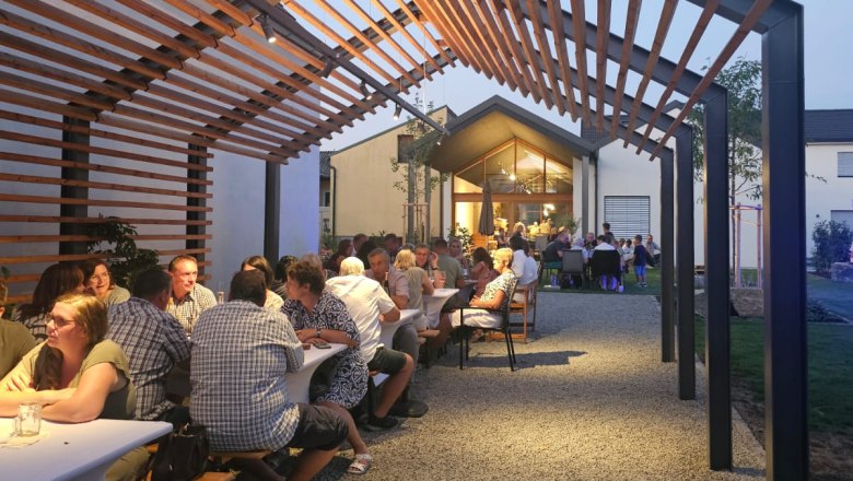 Menschen sitzen an langen Tischen unter einem Holzpergola bei Abendd&auml;mmerung.