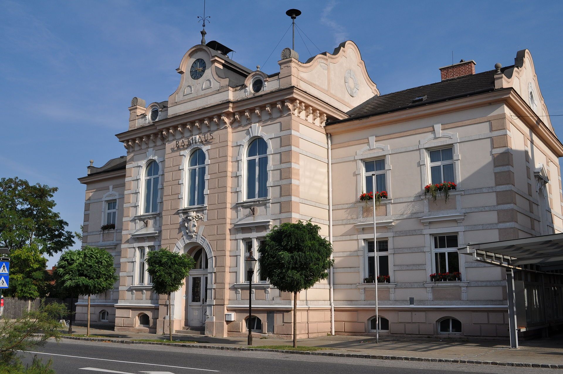Das Rathaus von Felixdorf, ein historisches Gebäude mit Uhrturm und verzierten Fenstern, umgeben von Bäumen und einer Straße im Vordergrund.