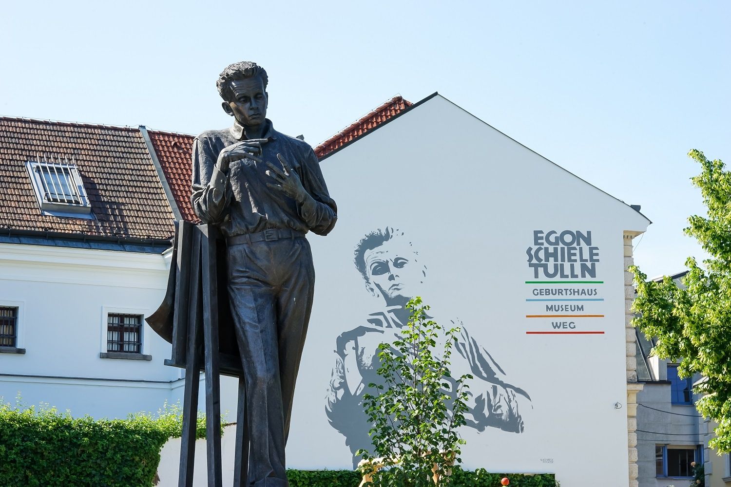 Statue von Egon Schiele vor einem Gebäude mit Wandgemälde und Schriftzug 'Egon Schiele Tulln'.