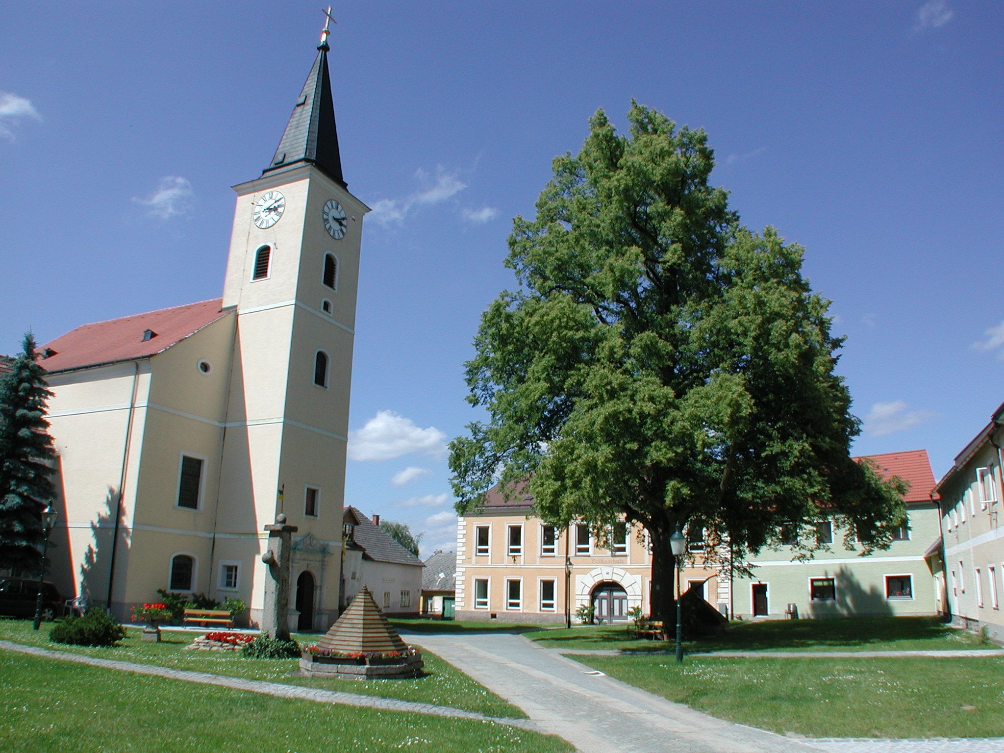 Kirche mit Turm und Uhr in einem Dorfplatz mit Bäumen und Gebäuden.