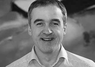 Schwarz-wei&szlig; Foto eines l&auml;chelnden Mannes in einem karierten Hemd.